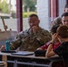 U.S. Army soldiers in multicam uniforms are seated with children in what appears to be a classroom or school setting, likely in the Netherlands based on the Dutch word 'Komodovaraan' (Komodo dragon) visible on a display board. The soldiers appear to be participating in an educational outreach or community engagement activity. The setting includes school desks, laptops, and educational materials.