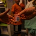 A person wearing a green military-style camouflage jacket is handing an embroidered American flag patch to another person whose open hand is extended to receive it. The patch features the standard U.S. flag design with red and white stripes and a blue field with stars, bordered in gold/orange. The scene appears to take place indoors with several people visible in the blurred background.