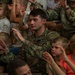U.S. Army soldiers in OCP (Operational Camouflage Pattern) uniforms are seated among a large group of young children, all raising their hands in what appears to be a group activity or interactive event. The soldiers, identifiable by 'U.S. ARMY' tape and what appears to be 101st Airborne Division patches, are actively participating alongside the children. The setting suggests a military family or community outreach event.