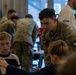 A U.S. Army soldier wearing an Airborne patch (101st Airborne Division) and identified by the name tape 'DAILEY' is leaning over and engaging with children at what appears to be a community or school event. Several other soldiers in camouflage uniforms are visible in the background, also interacting with children. The setting appears to be an indoor facility with large windows.