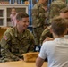 U.S. Army soldiers are visiting what appears to be a school or childcare facility, interacting with young children. The central soldier, identified by his name tape as 'TOBIN,' is smiling warmly while engaging with a child seated across from him. Multiple soldiers in OCP (Operational Camouflage Pattern) uniforms are present throughout the room.
