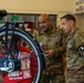 Two U.S. Army soldiers in OCP (Operational Camouflage Pattern) uniforms are examining a bicycle together in what appears to be a school or community center setting. The soldier on the left, identified by his name tape as 'FAZIO,' appears to be a senior NCO, while the soldier on the right wears a 101st Airborne Division (Ranger) patch. They appear to be participating in a community outreach or charity event involving bicycles.