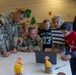 Two U.S. Army soldiers wearing camouflage uniforms with 101st Airborne Division patches are gathered around a laptop with three children at a white table. The setting appears to be an indoor community or school facility. The group appears to be engaged in a collaborative activity involving the laptop computer.