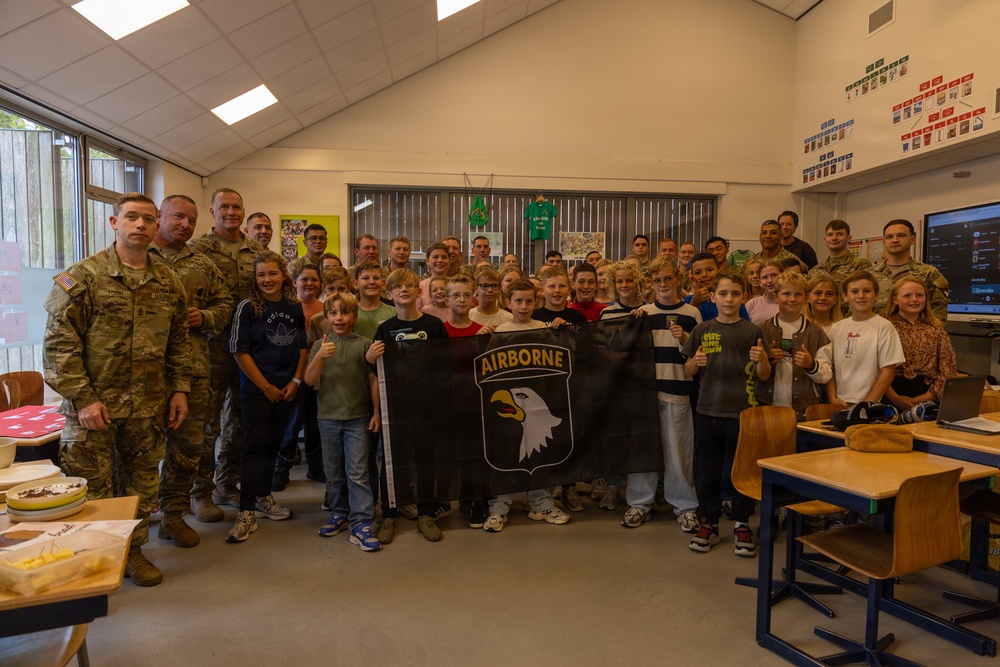 A group of U.S. Army soldiers from the 101st Airborne Division are posing with a classroom of children, likely during a school visit or community outreach event. Several children in the center are holding a black flag bearing the 101st Airborne Division insignia with an eagle and the word 'AIRBORNE'. The setting appears to be a European school classroom based on the furniture style and wall decorations.
