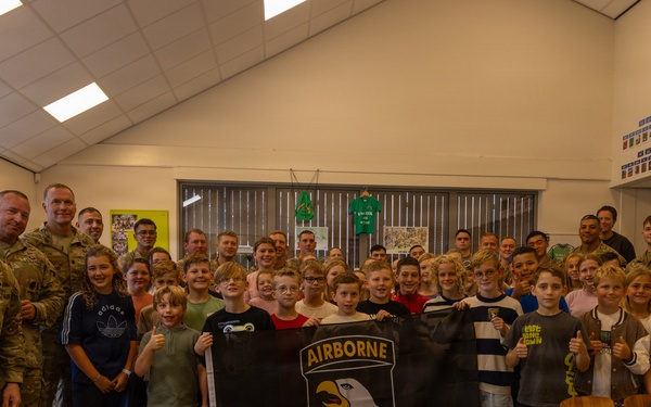 A group of U.S. Army soldiers from the 101st Airborne Division are posing with a classroom of children, likely during a school visit or community outreach event. Several children in the center are holding a black flag bearing the 101st Airborne Division insignia with an eagle and the word 'AIRBORNE'. The setting appears to be a European school classroom based on the furniture style and wall decorations.