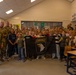 A group of U.S. Army soldiers from the 101st Airborne Division are posing with a classroom of children, likely during a school visit or community outreach event. Several children in the center are holding a black flag bearing the 101st Airborne Division insignia with an eagle and the word 'AIRBORNE'. The setting appears to be a European school classroom based on the furniture style and wall decorations.