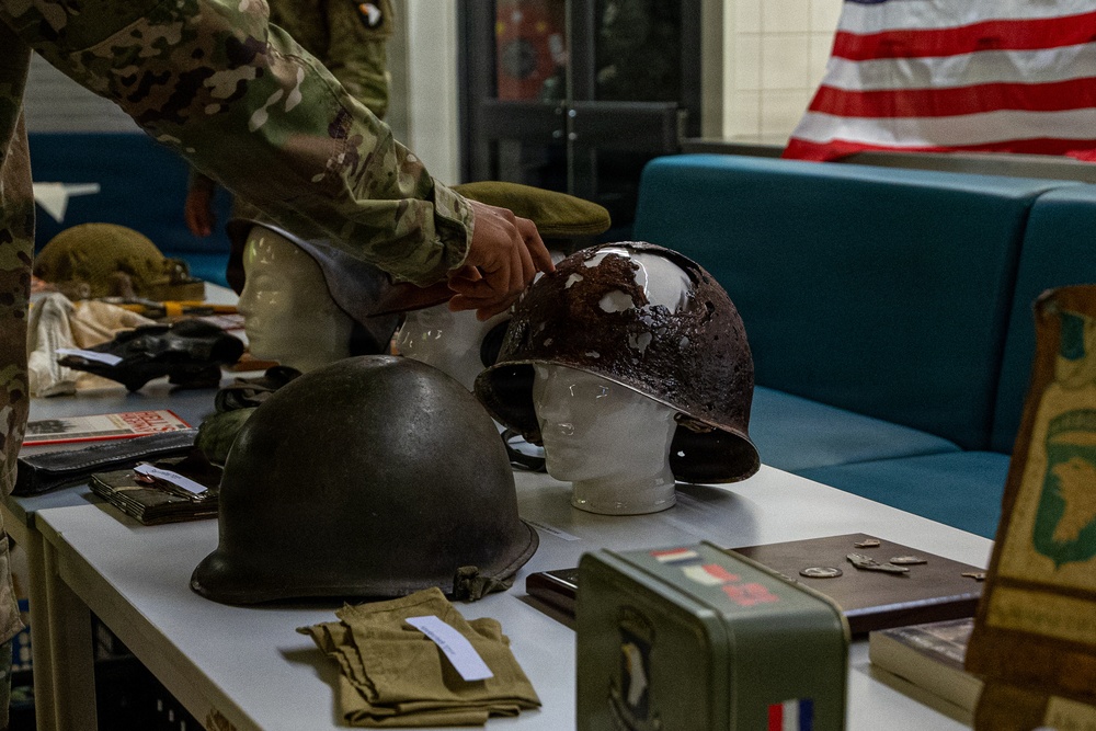 A military historical display table featuring WWII-era helmets and artifacts, with a person in modern U.S. Army camouflage uniform arranging items. The table holds multiple steel pot helmets, military accessories, medals, books, and a metal ammunition box. An American flag is visible in the background, suggesting this is an educational or commemorative exhibit.