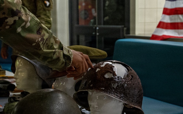 A military historical display table featuring WWII-era helmets and artifacts, with a person in modern U.S. Army camouflage uniform arranging items. The table holds multiple steel pot helmets, military accessories, medals, books, and a metal ammunition box. An American flag is visible in the background, suggesting this is an educational or commemorative exhibit.
