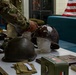 A military historical display table featuring WWII-era helmets and artifacts, with a person in modern U.S. Army camouflage uniform arranging items. The table holds multiple steel pot helmets, military accessories, medals, books, and a metal ammunition box. An American flag is visible in the background, suggesting this is an educational or commemorative exhibit.