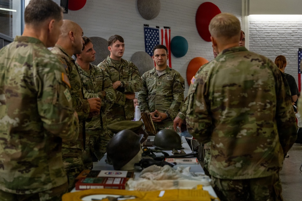 A group of U.S. Army soldiers in OCP (Operational Camouflage Pattern) uniforms are gathered around a table displaying historical military artifacts and equipment. The setting appears to be an indoor briefing or educational event, with an American flag visible on the wall in the background. The soldiers appear to be receiving a presentation or discussion about the displayed items.