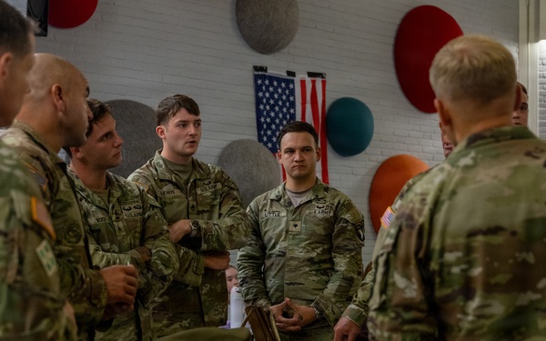 A group of U.S. Army soldiers in OCP (Operational Camouflage Pattern) uniforms are gathered around a table displaying historical military artifacts and equipment. The setting appears to be an indoor briefing or educational event, with an American flag visible on the wall in the background. The soldiers appear to be receiving a presentation or discussion about the displayed items.