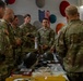 A group of U.S. Army soldiers in OCP (Operational Camouflage Pattern) uniforms are gathered around a table displaying historical military artifacts and equipment. The setting appears to be an indoor briefing or educational event, with an American flag visible on the wall in the background. The soldiers appear to be receiving a presentation or discussion about the displayed items.
