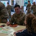 A U.S. Army soldier named Jeong, wearing the rank of Sergeant, is seated at a table smiling and interacting with a curly-haired civilian, likely a student or child. The setting appears to be a school or community center where multiple soldiers in OCP (Operational Camouflage Pattern) uniforms are engaging with civilians and children. The event appears to be a community outreach or school visit program.