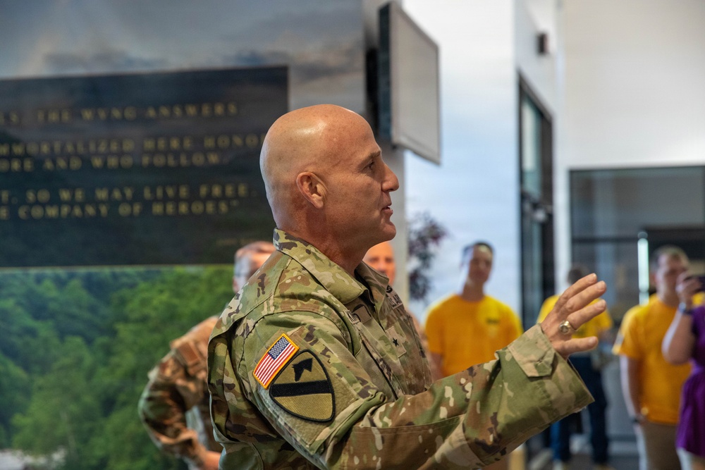 A U.S. Army officer in MultiCam/OCP camouflage uniform is speaking or presenting to a group of civilians, many wearing yellow shirts, in what appears to be a memorial or museum setting. The officer wears a 1st Cavalry Division patch and a U.S. flag patch on the left sleeve, with a single star rank insignia visible, indicating the rank of Brigadier General. A dark memorial wall with inscribed text is visible in the background.