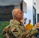 A U.S. Army officer in MultiCam/OCP camouflage uniform is speaking or presenting to a group of civilians, many wearing yellow shirts, in what appears to be a memorial or museum setting. The officer wears a 1st Cavalry Division patch and a U.S. flag patch on the left sleeve, with a single star rank insignia visible, indicating the rank of Brigadier General. A dark memorial wall with inscribed text is visible in the background.
