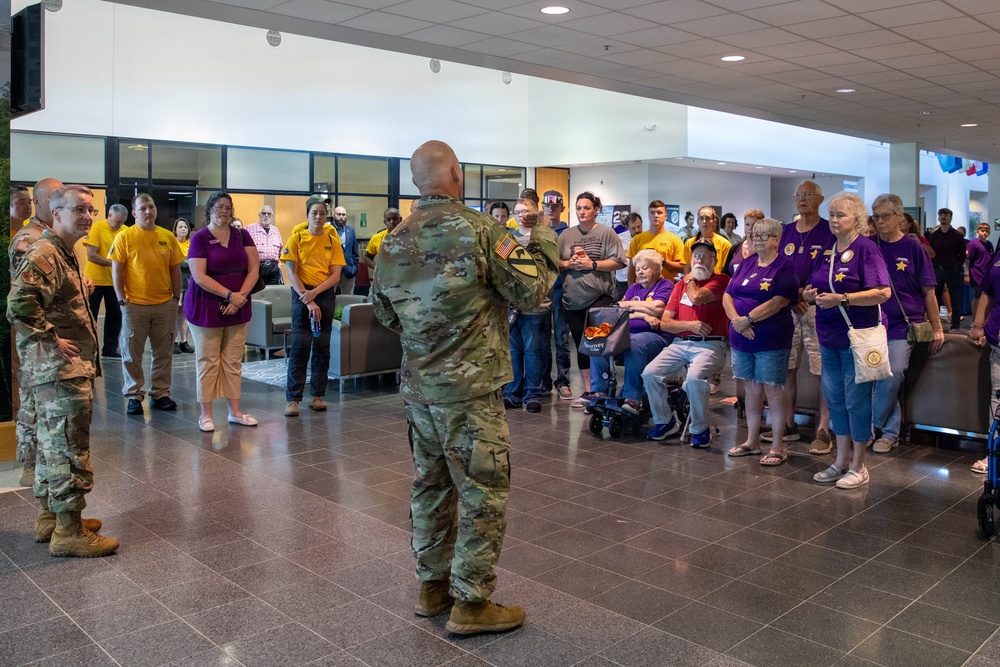 A military briefing or welcome ceremony is taking place in a modern building lobby, where two soldiers in Army Combat Uniforms (ACU/OCP camouflage) are addressing a large group of civilians. The civilians are wearing coordinated yellow and purple t-shirts, suggesting they are part of an organized group, possibly a veterans support organization or Gold Star family event. One soldier faces the crowd while another stands to the side, indicating a formal address or presentation.
