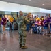 A military briefing or welcome ceremony is taking place in a modern building lobby, where two soldiers in Army Combat Uniforms (ACU/OCP camouflage) are addressing a large group of civilians. The civilians are wearing coordinated yellow and purple t-shirts, suggesting they are part of an organized group, possibly a veterans support organization or Gold Star family event. One soldier faces the crowd while another stands to the side, indicating a formal address or presentation.