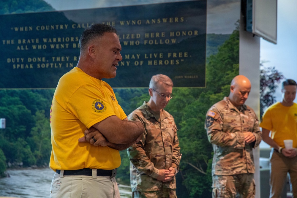 A group of individuals, including two military personnel in camouflage uniforms and civilians in yellow shirts, are gathered in what appears to be a solemn ceremony or moment of reflection. A large commemorative plaque or mural is visible in the background referencing the West Virginia National Guard (WVNG) with an inspirational quote. The setting suggests a memorial or dedication event.