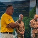 A group of individuals, including two military personnel in camouflage uniforms and civilians in yellow shirts, are gathered in what appears to be a solemn ceremony or moment of reflection. A large commemorative plaque or mural is visible in the background referencing the West Virginia National Guard (WVNG) with an inspirational quote. The setting suggests a memorial or dedication event.