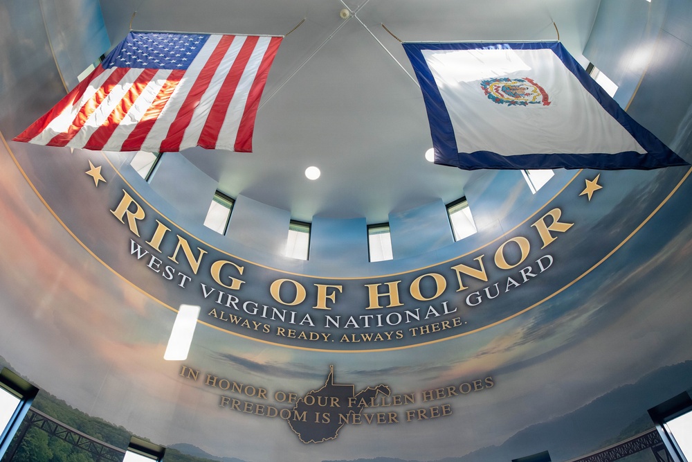 The image shows the interior of the West Virginia National Guard 'Ring of Honor' memorial, photographed from a low angle looking upward at a curved ceiling. Two flags — the United States flag and the West Virginia state flag — are displayed prominently. The installation features inspirational text honoring fallen military heroes.