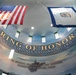 The image shows the interior of the West Virginia National Guard 'Ring of Honor' memorial, photographed from a low angle looking upward at a curved ceiling. Two flags — the United States flag and the West Virginia state flag — are displayed prominently. The installation features inspirational text honoring fallen military heroes.