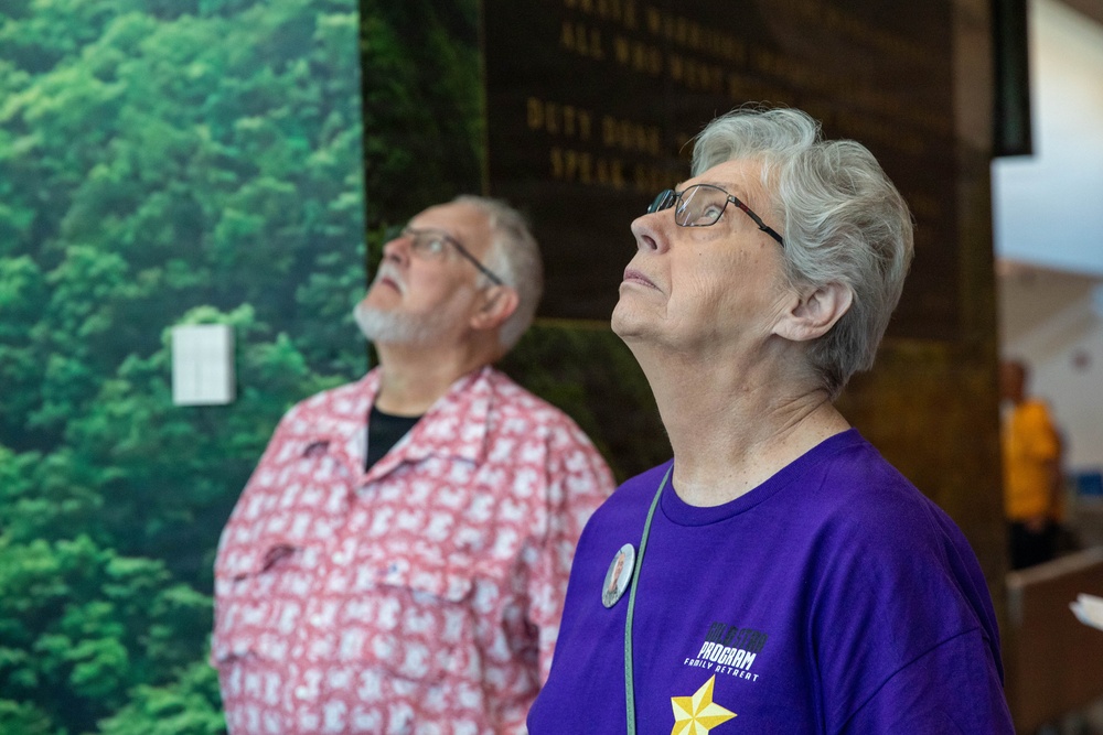 Two older adults are looking upward in what appears to be a museum or memorial exhibit. The woman in the foreground wears a purple t-shirt reading 'Family Retreat Program' with a gold star graphic and a button pin, while the man behind her wears a patterned red shirt. They appear to be viewing something above them, possibly a display or presentation.