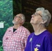 Two older adults are looking upward in what appears to be a museum or memorial exhibit. The woman in the foreground wears a purple t-shirt reading 'Family Retreat Program' with a gold star graphic and a button pin, while the man behind her wears a patterned red shirt. They appear to be viewing something above them, possibly a display or presentation.