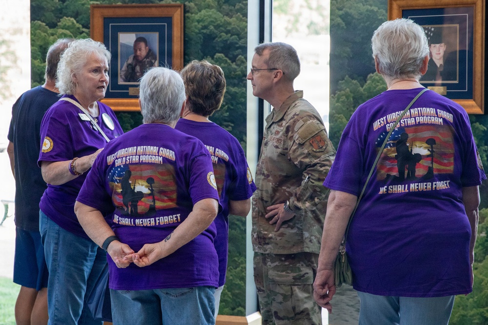 A group of people wearing purple 'West Virginia National Guard Gold Star Program' t-shirts with the motto 'We Shall Never Forget' are gathered in what appears to be a memorial or official building. They are conversing with a military service member in camouflage uniform. Framed portraits of military personnel are visible on the wall in the background.