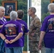 A group of people wearing purple 'West Virginia National Guard Gold Star Program' t-shirts with the motto 'We Shall Never Forget' are gathered in what appears to be a memorial or official building. They are conversing with a military service member in camouflage uniform. Framed portraits of military personnel are visible on the wall in the background.