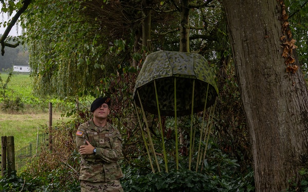 A U.S. Army soldier in modern MultiCam (OCP) uniform stands with arms crossed beside a memorial monument dedicated to the 101st Airborne Division, commemorating their landing in September 1944. The monument features a decorative parachute sculpture in camouflage colors and a plaque with Dutch and American flags. The setting appears to be a rural area in the Netherlands, consistent with Operation Market Garden commemorative sites.