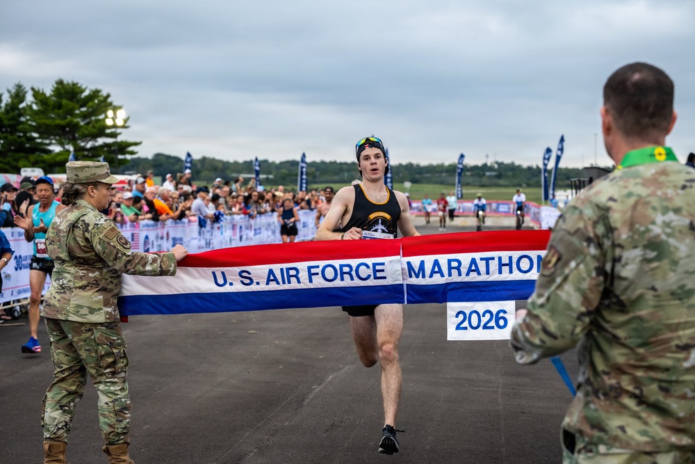 A runner is crossing the finish line at the U.S. Air Force Marathon 2026, breaking through a red, white, and blue banner held by two military personnel in camouflage uniforms. The event appears to be taking place on a tarmac or airfield with a large crowd of spectators visible in the background. The runner, wearing a black sleeveless top and a cap, appears to be the first-place finisher.