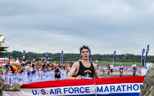 A runner is crossing the finish line at the U.S. Air Force Marathon 2026, breaking through a red, white, and blue banner held by two military personnel in camouflage uniforms. The event appears to be taking place on a tarmac or airfield with a large crowd of spectators visible in the background. The runner, wearing a black sleeveless top and a cap, appears to be the first-place finisher.