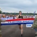 A runner is crossing the finish line at the U.S. Air Force Marathon 2026, breaking through a red, white, and blue banner held by two military personnel in camouflage uniforms. The event appears to be taking place on a tarmac or airfield with a large crowd of spectators visible in the background. The runner, wearing a black sleeveless top and a cap, appears to be the first-place finisher.