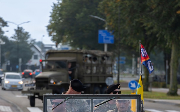 A WWII-era Willys MB jeep marked '508' and '82 AB' (82nd Airborne) participates in a commemorative military convoy on a Dutch road, identifiable by the Dutch license plate 'GR-48-ZK' and blue Dutch road signs. The jeep carries several occupants in military attire and displays an 82nd Airborne Division flag along with a yellow flag. A period military truck follows behind in the convoy.