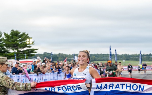A female runner is crossing the finish line at the US Air Force Marathon, breaking through a red, white, and blue finisher's tape. She is smiling broadly, suggesting she is the first female finisher or overall winner. The event is held outdoors on a paved surface with a large crowd of spectators visible in the background.