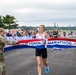 A female runner is crossing the finish line at the US Air Force Marathon, breaking through a red, white, and blue finisher's tape. She is smiling broadly, suggesting she is the first female finisher or overall winner. The event is held outdoors on a paved surface with a large crowd of spectators visible in the background.