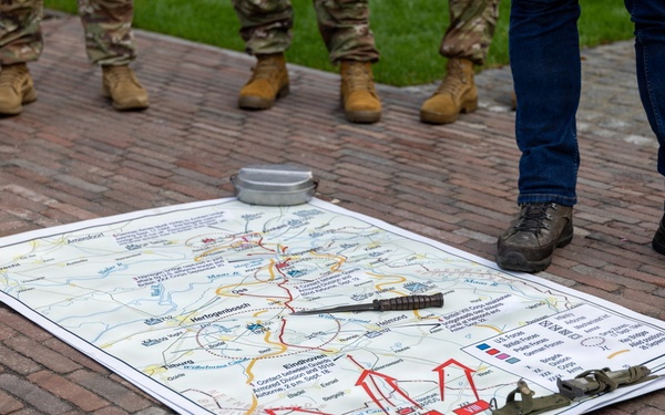 A military tactical map is laid out on a brick surface, weighted down at the corners with military equipment. The map depicts the Netherlands region including cities such as Eindhoven, Tilburg, and 's-Hertogenbosch, with red arrows indicating troop movements consistent with Operation Market Garden (WWII). Several individuals in military boots and one civilian are standing around the map, suggesting a battlefield staff ride or historical briefing.