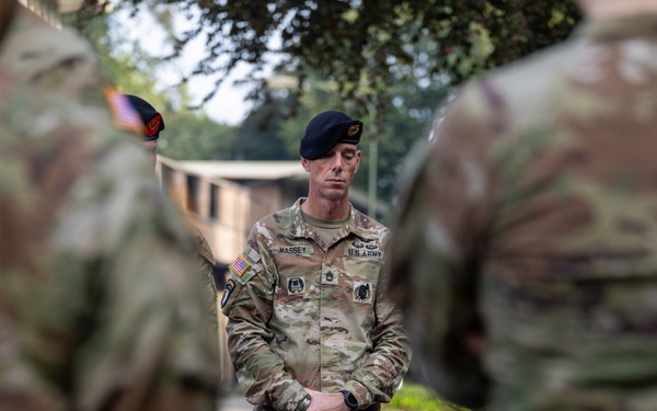 A U.S. Army soldier identified by name tape as 'MASSEY' stands at attention or in a relaxed parade rest position, wearing OCP (Operational Camouflage Pattern) uniform and a black beret. He appears to be addressing or observing other soldiers, framed between two out-of-focus uniformed personnel in the foreground. On the ground in front of him are materials including what appears to be a map booklet labeled 'TRACES OF WAR.'