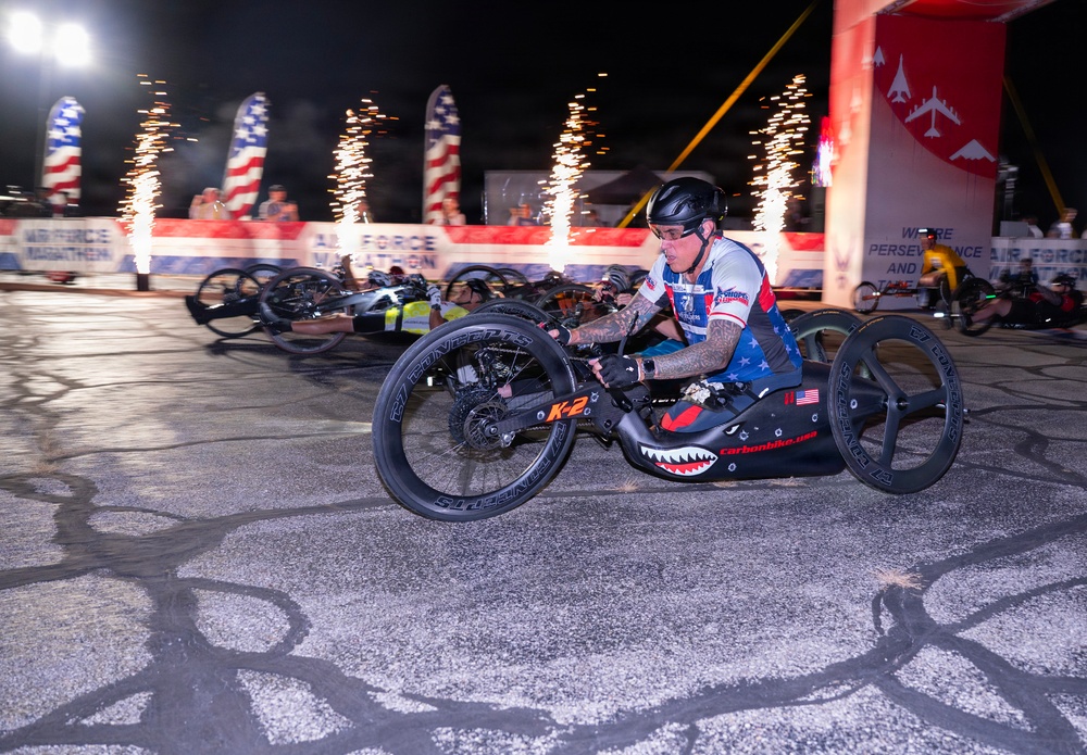 A nighttime hand-cycling race is underway at what appears to be the Air Force Marathon, with multiple athletes competing on specialized hand-powered racing cycles. The lead athlete is prominently featured in a patriotic red, white, and blue jersey, riding a carbon fiber hand cycle branded 'carbonbike.usa' with a shark mouth design. Pyrotechnic fireworks and American flag banners illuminate the dramatic race start atmosphere in the background.