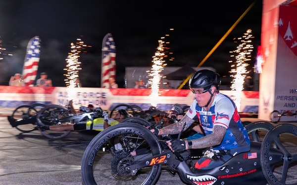A nighttime hand-cycling race is underway at what appears to be the Air Force Marathon, with multiple athletes competing on specialized hand-powered racing cycles. The lead athlete is prominently featured in a patriotic red, white, and blue jersey, riding a carbon fiber hand cycle branded 'carbonbike.usa' with a shark mouth design. Pyrotechnic fireworks and American flag banners illuminate the dramatic race start atmosphere in the background.