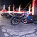 A nighttime hand-cycling race is underway at what appears to be the Air Force Marathon, with multiple athletes competing on specialized hand-powered racing cycles. The lead athlete is prominently featured in a patriotic red, white, and blue jersey, riding a carbon fiber hand cycle branded 'carbonbike.usa' with a shark mouth design. Pyrotechnic fireworks and American flag banners illuminate the dramatic race start atmosphere in the background.