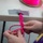 A person is crafting a paracord bracelet using neon pink and multicolored cord. The work-in-progress bracelet is anchored to the edge of a table using duct tape and a clip or clasp. A spool of neon pink cord is visible in the background, suggesting an ongoing crafting session.