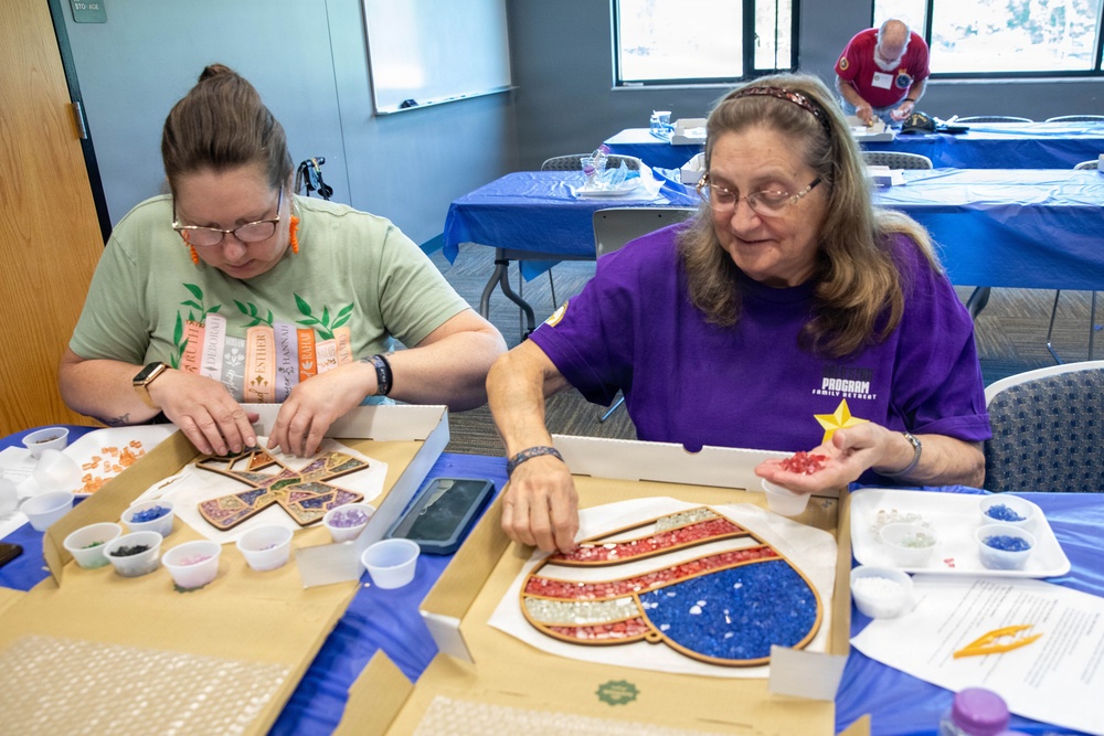 Two women are seated at a table engaged in a mosaic craft activity, applying small colored glass or stone pieces to decorative wooden shapes. The woman on the left works on a cross-shaped mosaic, while the woman on the right works on a heart-shaped mosaic with red, white, and blue colors. A third participant is visible in the background working at a separate table in what appears to be a community center or classroom setting.