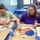 Two women are seated at a table engaged in a mosaic craft activity, applying small colored glass or stone pieces to decorative wooden shapes. The woman on the left works on a cross-shaped mosaic, while the woman on the right works on a heart-shaped mosaic with red, white, and blue colors. A third participant is visible in the background working at a separate table in what appears to be a community center or classroom setting.