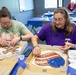 Two women are seated at a table engaged in a mosaic craft activity, applying small colored glass or stone pieces to decorative wooden shapes. The woman on the left works on a cross-shaped mosaic, while the woman on the right works on a heart-shaped mosaic with red, white, and blue colors. A third participant is visible in the background working at a separate table in what appears to be a community center or classroom setting.