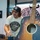 A man wearing a 'Commemorate Through Choice' t-shirt and matching snapback cap stands behind a table next to a signed acoustic guitar. The guitar features multiple white autographs on its dark body, and a matching 'Commemorate' banner is visible in the background. The setting appears to be an indoor event space, possibly a meet-and-greet or signing event.