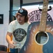 A man wearing a 'Commemorate Through Choice' t-shirt and matching snapback cap stands behind a table next to a signed acoustic guitar. The guitar features multiple white autographs on its dark body, and a matching 'Commemorate' banner is visible in the background. The setting appears to be an indoor event space, possibly a meet-and-greet or signing event.