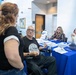 A group of people are gathered around a blue-draped information table in what appears to be a public building or community center. An older man in a wheelchair wearing a black commemorative t-shirt is engaged in conversation with a woman standing with her back to the camera. Two women seated behind the table appear to be staffing the booth, with one smiling.