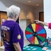 Two individuals are interacting at what appears to be an informational booth or event table. A woman in a pink and black shirt wearing a patriotic headband is spinning a colorful prize wheel for an older person wearing a purple West Virginia National Guard Gold Star Program t-shirt. The setting appears to be an indoor facility, likely a community or veterans event.
