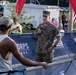A U.S. Marine Corps officer in camouflage uniform is speaking into a microphone at what appears to be a Marine Corps Marathon event at New River. He is addressing a crowd near a race barrier, with banners reading 'Marine Corps' and 'New River' visible in the background. American and red flags are visible, along with spectators and other Marines in the background.